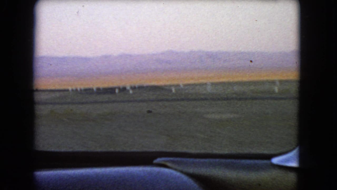A 1966 Nevada desert road view through the car window, with white roadside posts crossing the foreground and mountains in haze.