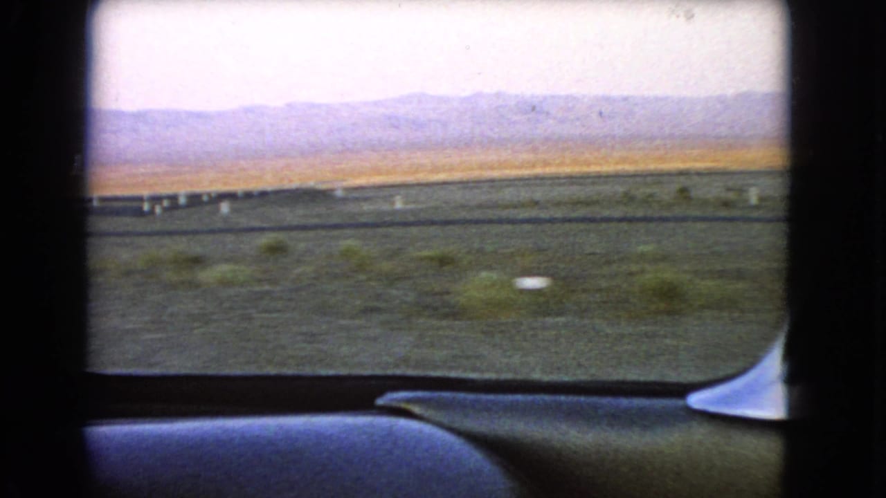 End frame from a 1966 Nevada car-window shot, showing passing desert shrubs, marker posts, open plain, and distant mountain silhouettes.