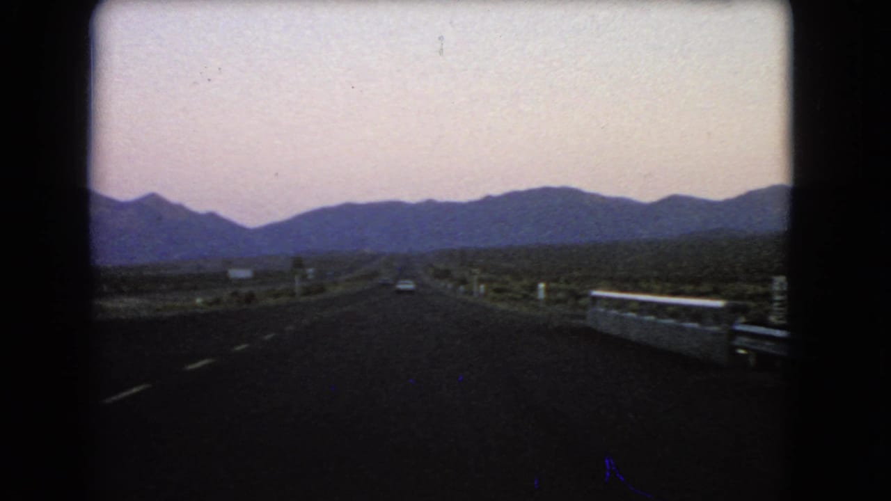 Desert highway stretches toward distant mountains under a pale sky.