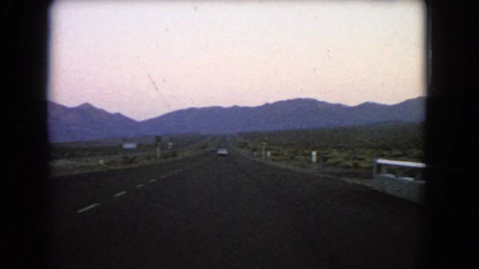 Wide 1966 Nevada road view with a dark highway receding toward mountain silhouettes, a small vehicle ahead, roadside posts, desert scrub, and visible 8mm frame edges.