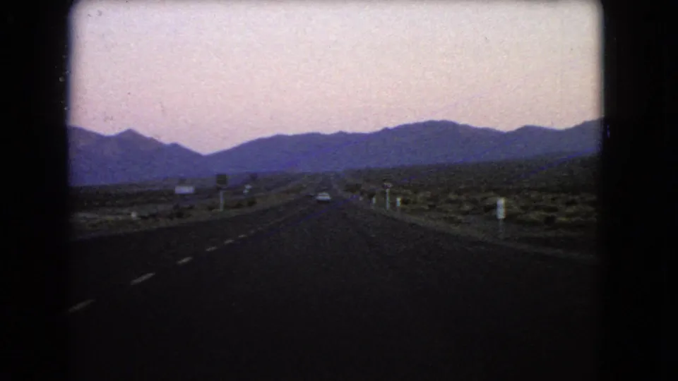 Twilight road scene from the 1966 Nevada 8mm clip, showing the highway receding into desert terrain with mountain silhouettes, roadside markers, and a soft pink-blue sky.
