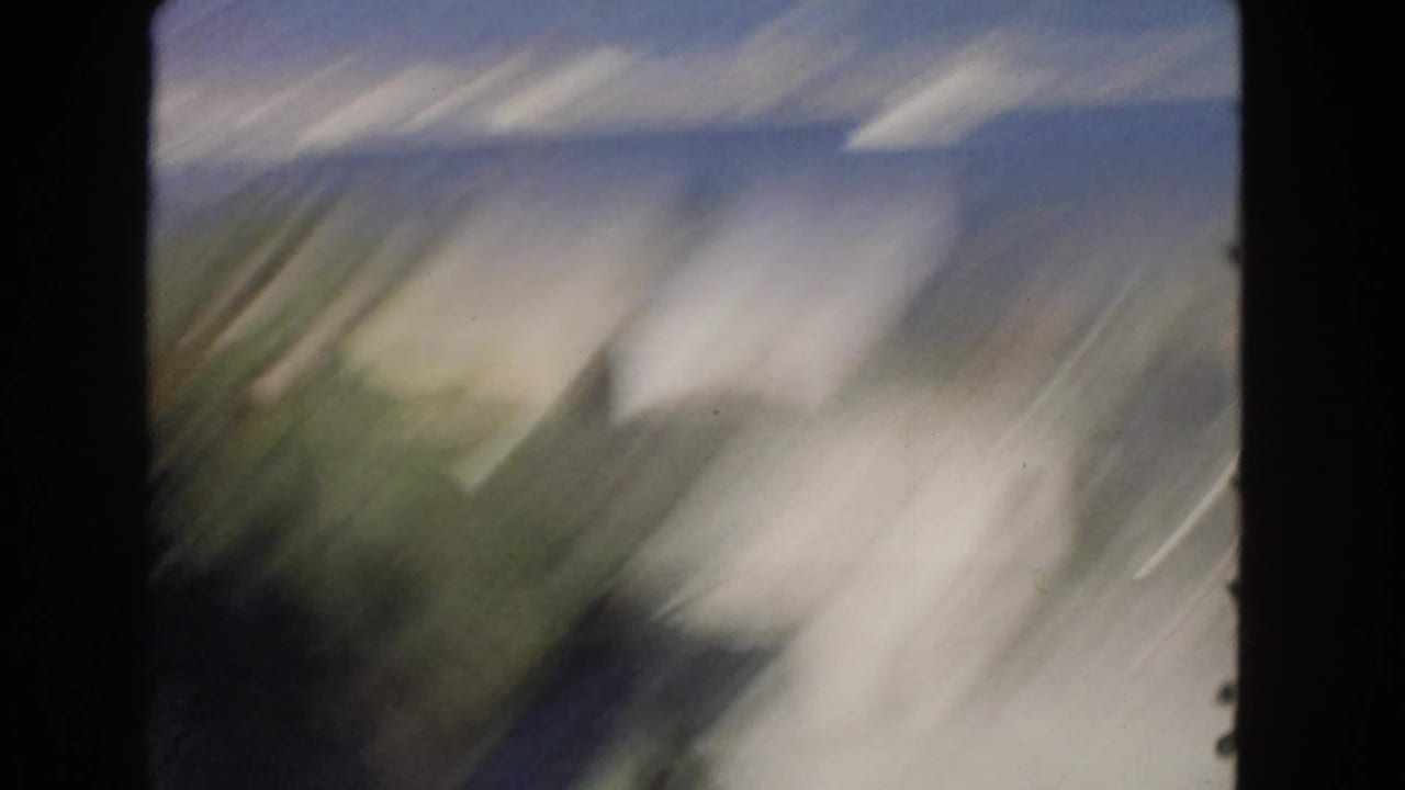 Blurred handheld 8mm movement over a San Pedro street area with pale buildings, road surface, and indistinct foreground shapes.