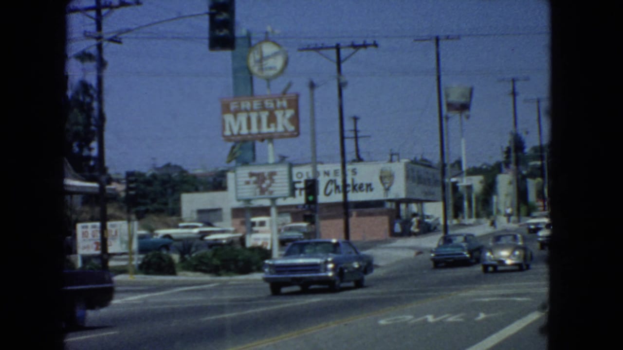 Busy street with cars passing a fried chicken restaurant and large Fresh Milk sign.
