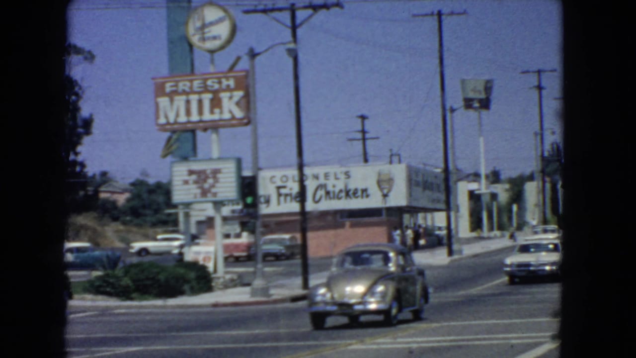 A Volkswagen Beetle moves through a 1966 San Pedro street scene near a Fresh Milk sign and Colonel's Kentucky Fried Chicken storefront.