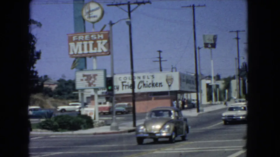 A Volkswagen Beetle moves through a 1966 San Pedro street scene near a Fresh Milk sign and Colonel's Kentucky Fried Chicken storefront.