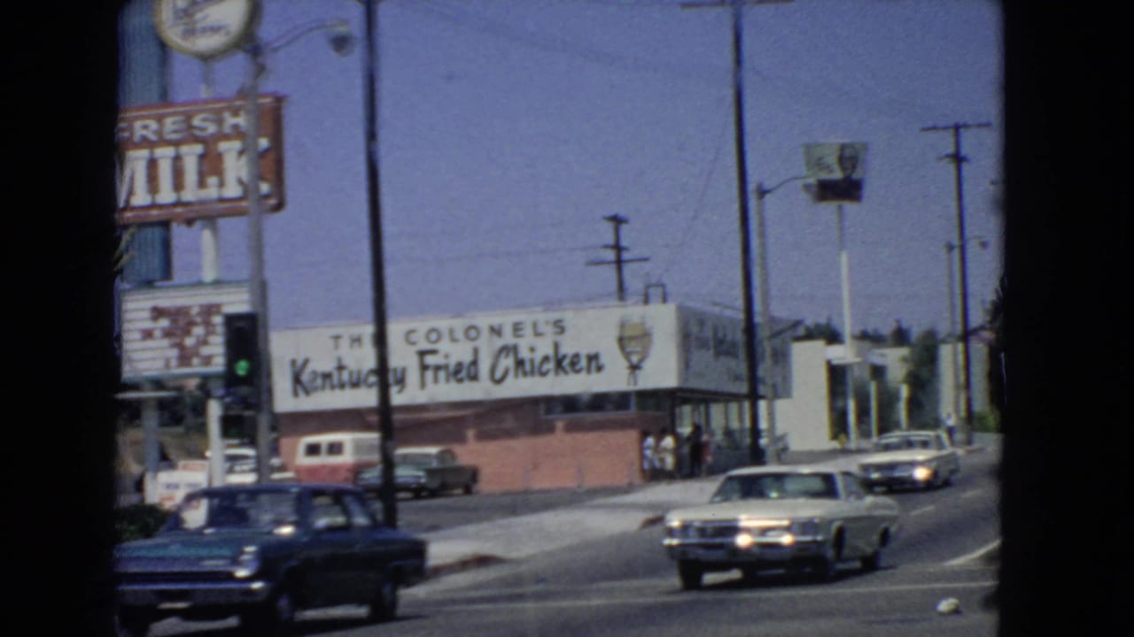 1966 San Pedro street view showing a Colonel's Kentucky Fried Chicken restaurant, a roadside milk sign, utility poles, and mid-1960s cars moving along the road.