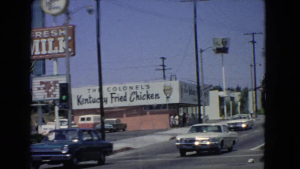 1966 San Pedro street view showing a Colonel's Kentucky Fried Chicken restaurant, a roadside milk sign, utility poles, and mid-1960s cars moving along the road.
