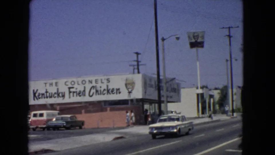1966 San Pedro footage frame showing the Kentucky Fried Chicken exterior, a parked red-and-white van, pedestrians on the sidewalk, and a light-colored car approaching on the road.