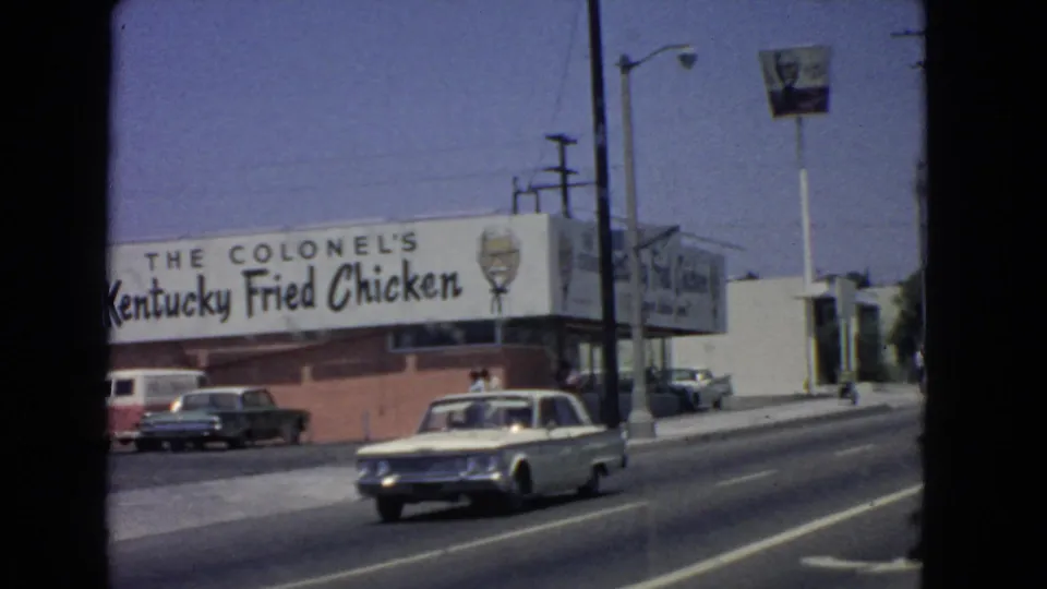 1966 San Pedro street frame with a light-colored sedan passing the Colonel's Kentucky Fried Chicken restaurant, a parked van, pedestrians, street markings, and roadside signs.