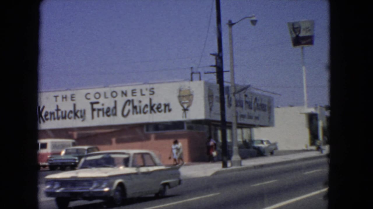 Street view passing The Colonel's Kentucky Fried Chicken building with cars and pedestrians.