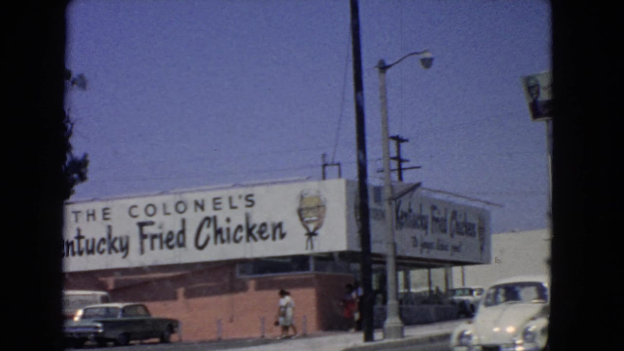 Kentucky Fried Chicken storefront seen from the street with cars, pedestrians, and bucket sign.