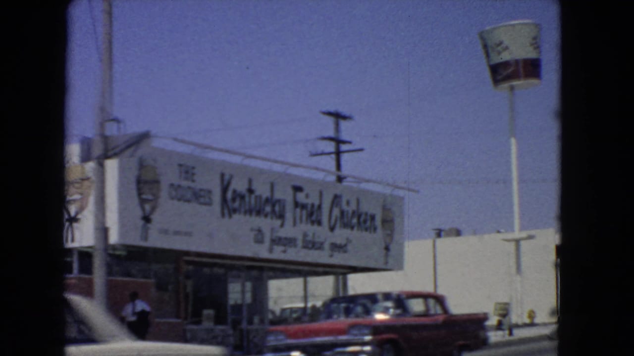 Drive-by view of a Kentucky Fried Chicken storefront sign with a red car passing in front.