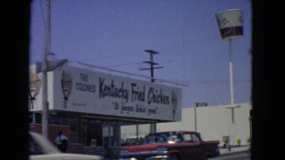 1966 San Pedro drive-by frame showing a Kentucky Fried Chicken sign, bucket logo, red period car, and roadside utility poles.
