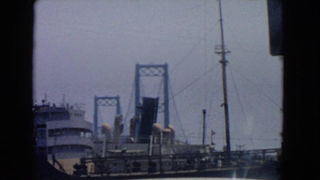 1966 San Pedro harbor view showing a ship beside bridge towers, waterfront railings, utility poles, and hazy blue sky.