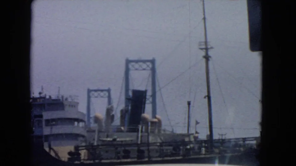 1966 San Pedro harbor view showing a ship beside bridge towers, waterfront railings, utility poles, and hazy blue sky.