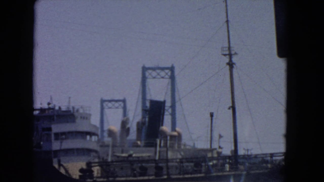 San Pedro port-area frame with a ship at left, bridge towers behind it, roadside poles, railings, and vehicle-edge framing.