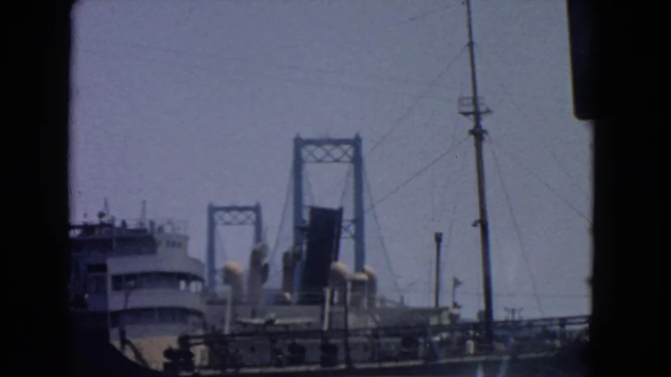 San Pedro port-area frame with a ship at left, bridge towers behind it, roadside poles, railings, and vehicle-edge framing.