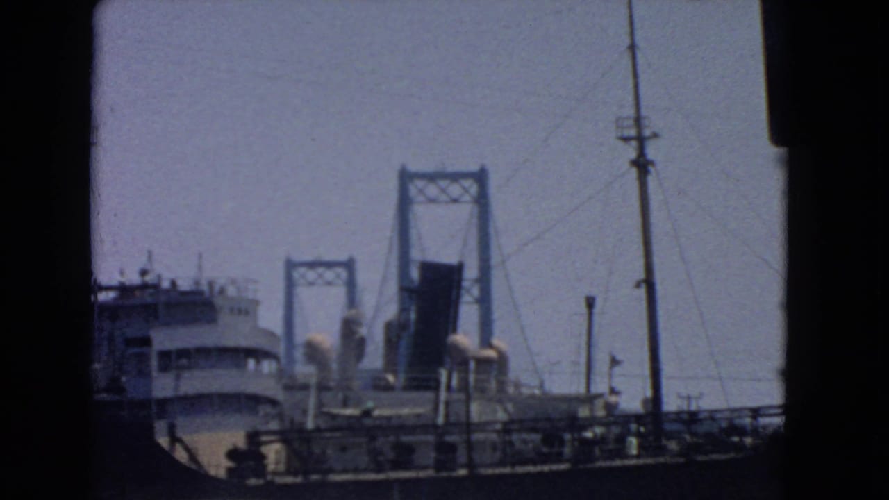 1966 San Pedro harbor view with a large ship in the foreground and a suspension bridge rising behind it under hazy light.