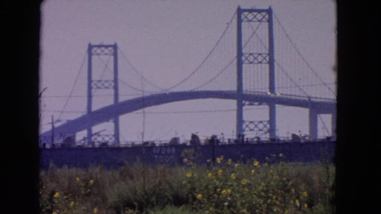 1966 San Pedro home-movie frame showing the bridge span and towers beyond grass under a hazy sky.