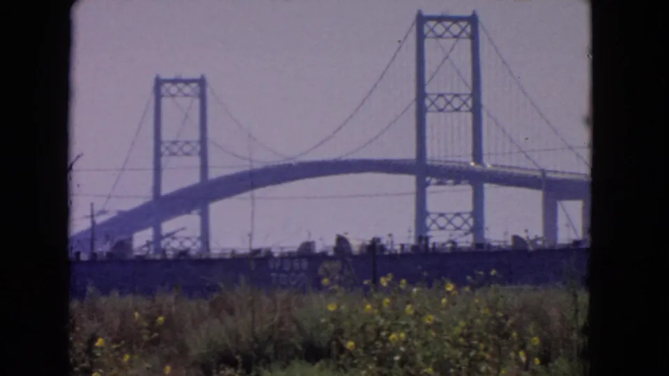 1966 San Pedro home-movie frame showing the bridge span and towers beyond grass under a hazy sky.