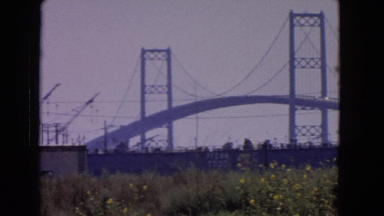 1966 San Pedro shoreline view of a bridge under construction, with towers, cables, grass, and small industrial shapes near the foreground.