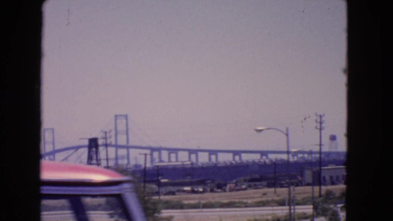 1966 San Pedro roadside view of the Vincent Thomas Bridge, industrial structures, poles, and a red vehicle passing low across the foreground.