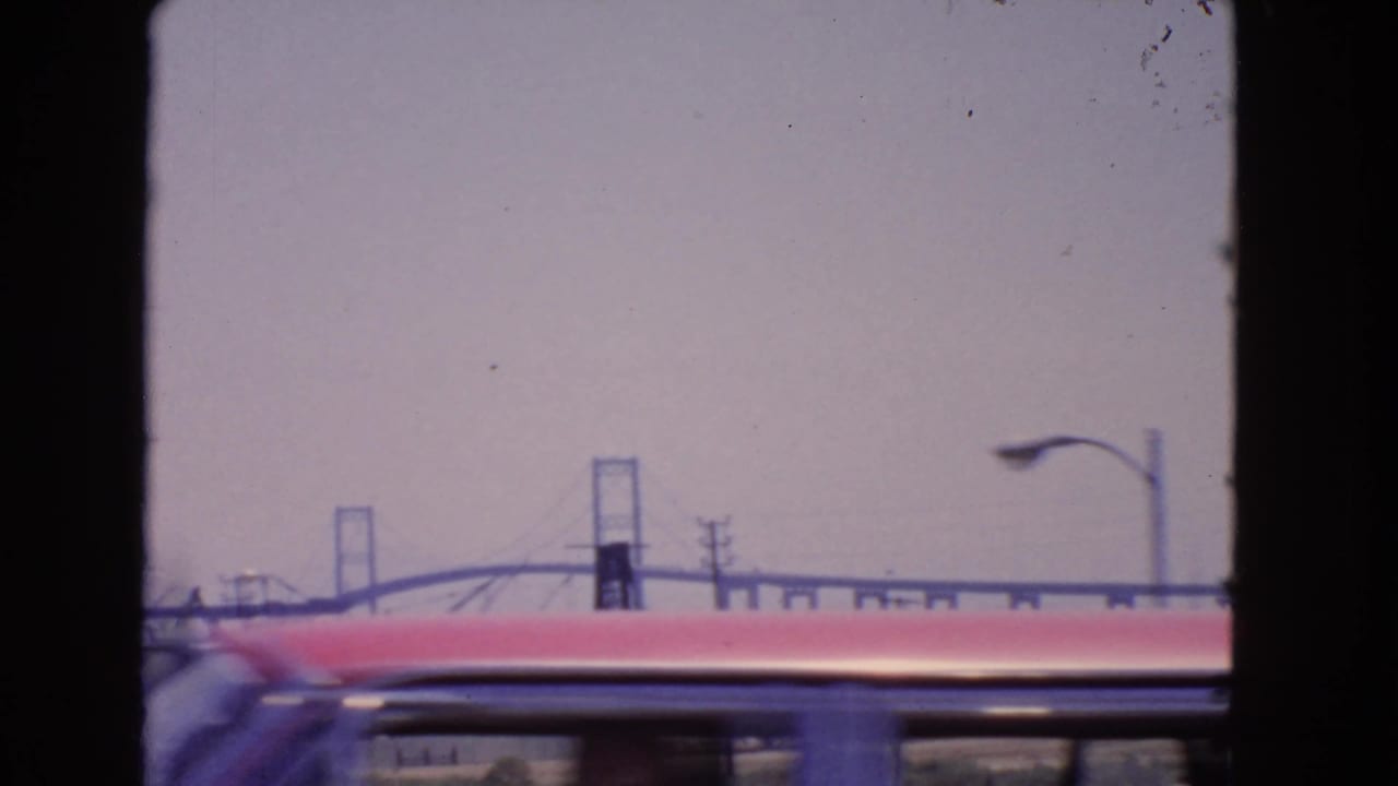1966 San Pedro frame with a red vehicle passing across the foreground while the Vincent Thomas Bridge, streetlight, and poles sit behind it.