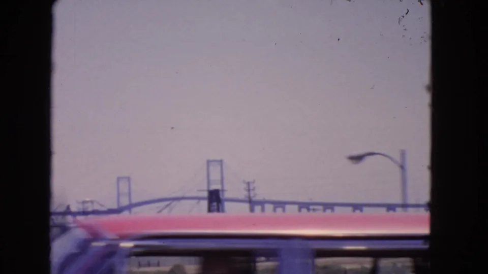 1966 San Pedro frame with a red vehicle passing across the foreground while the Vincent Thomas Bridge, streetlight, and poles sit behind it.