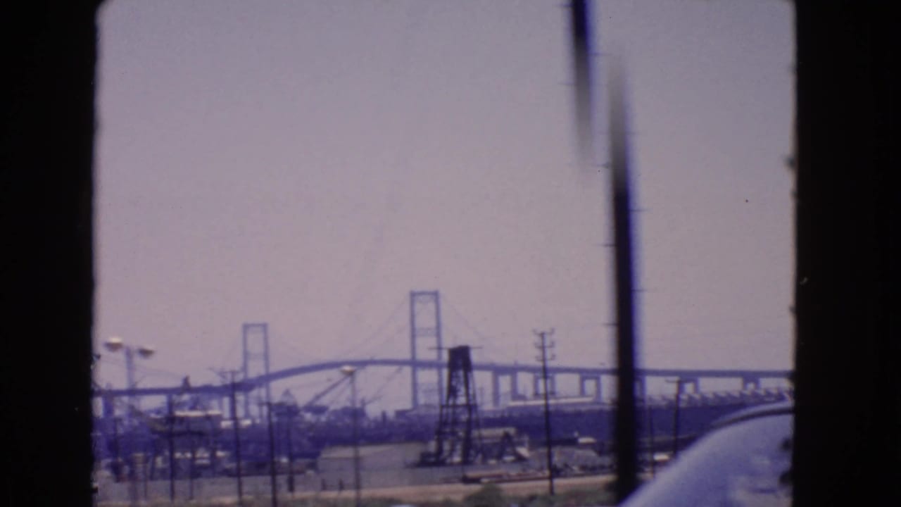 1966 San Pedro view of an industrial bridge construction area framed by a window, with cranes, power lines, and road traffic below.
