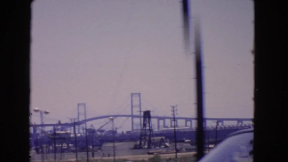1966 San Pedro view of an industrial bridge construction area framed by a window, with cranes, power lines, and road traffic below.