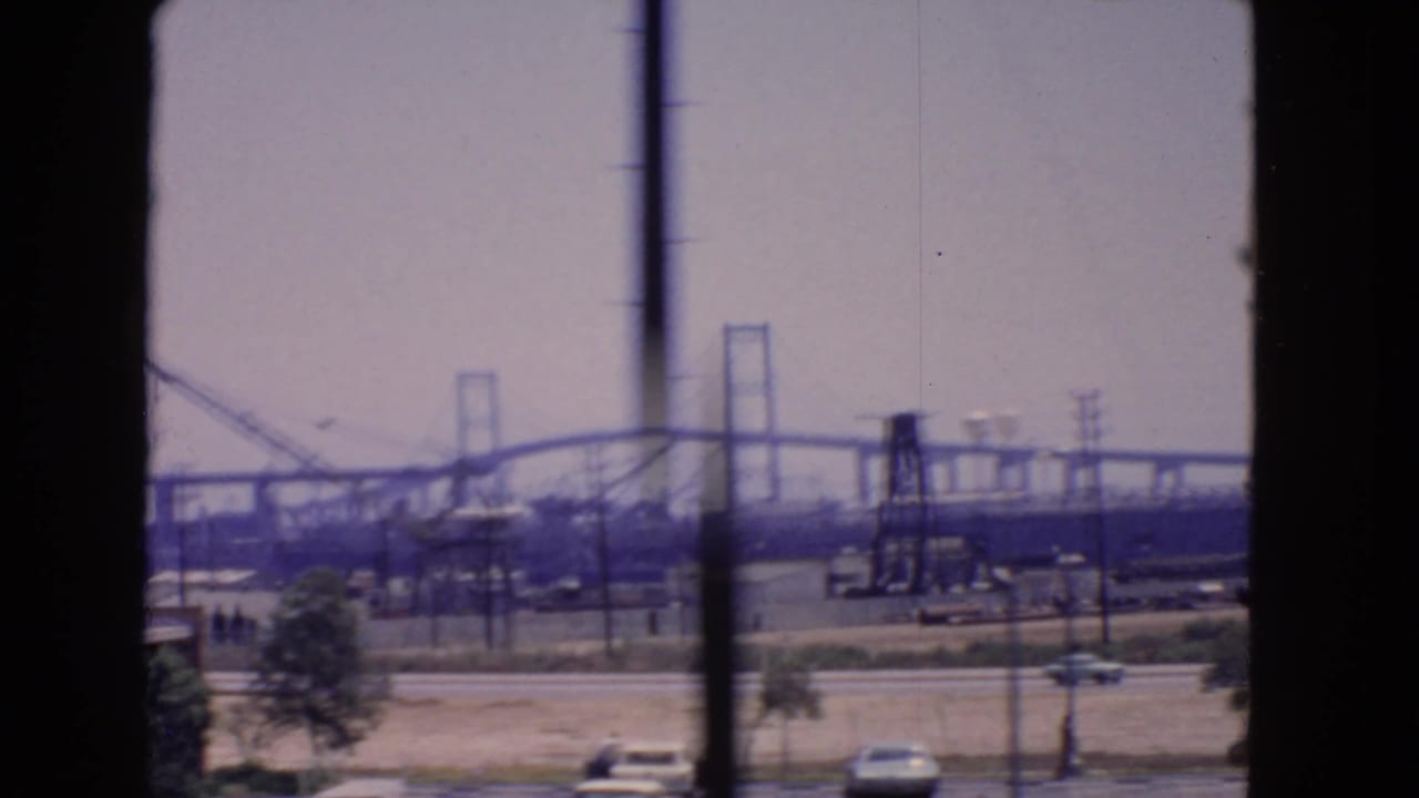 Distant bridge span with cranes, utility poles, roadside cars, and a dark vertical window frame.