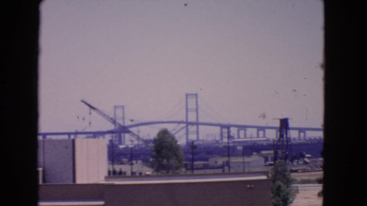 San Pedro, California, 1966 view of a bridge construction area beyond low industrial buildings, trees, utility lines, and passing cars.