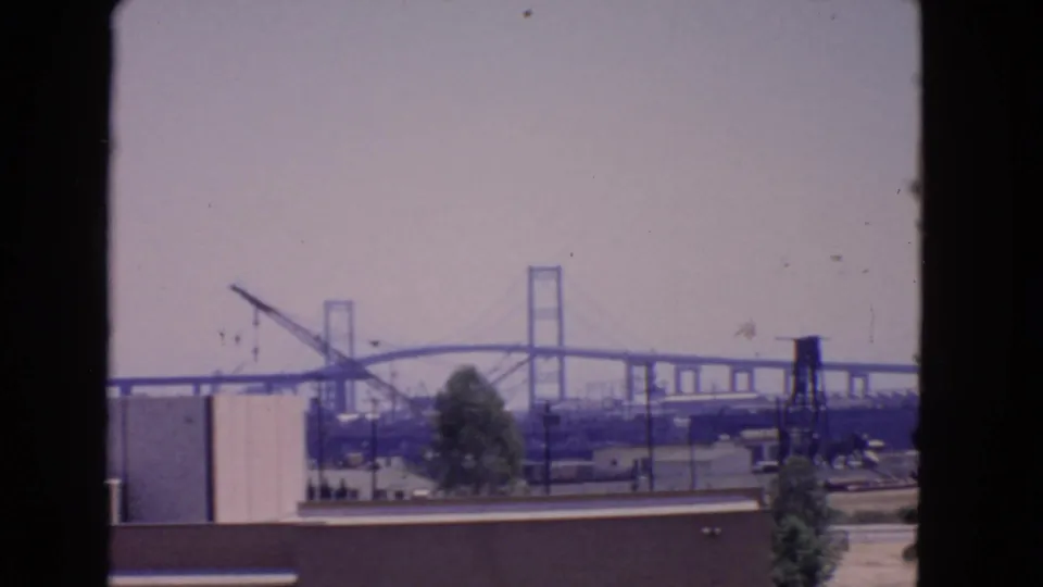 San Pedro, California, 1966 view of a bridge construction area beyond low industrial buildings, trees, utility lines, and passing cars.