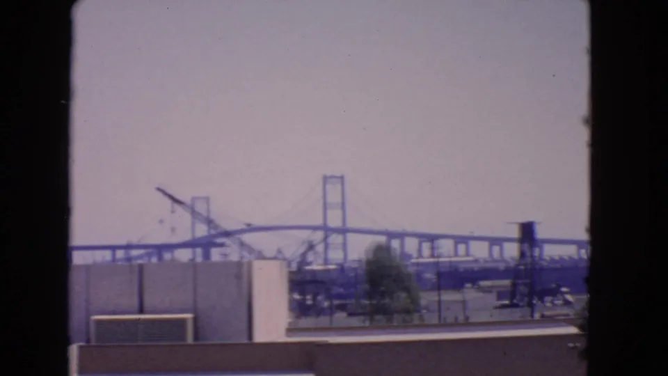 1966 San Pedro view with a distant suspension bridge, waterfront structures, rooftops, and soft 8mm color fade.