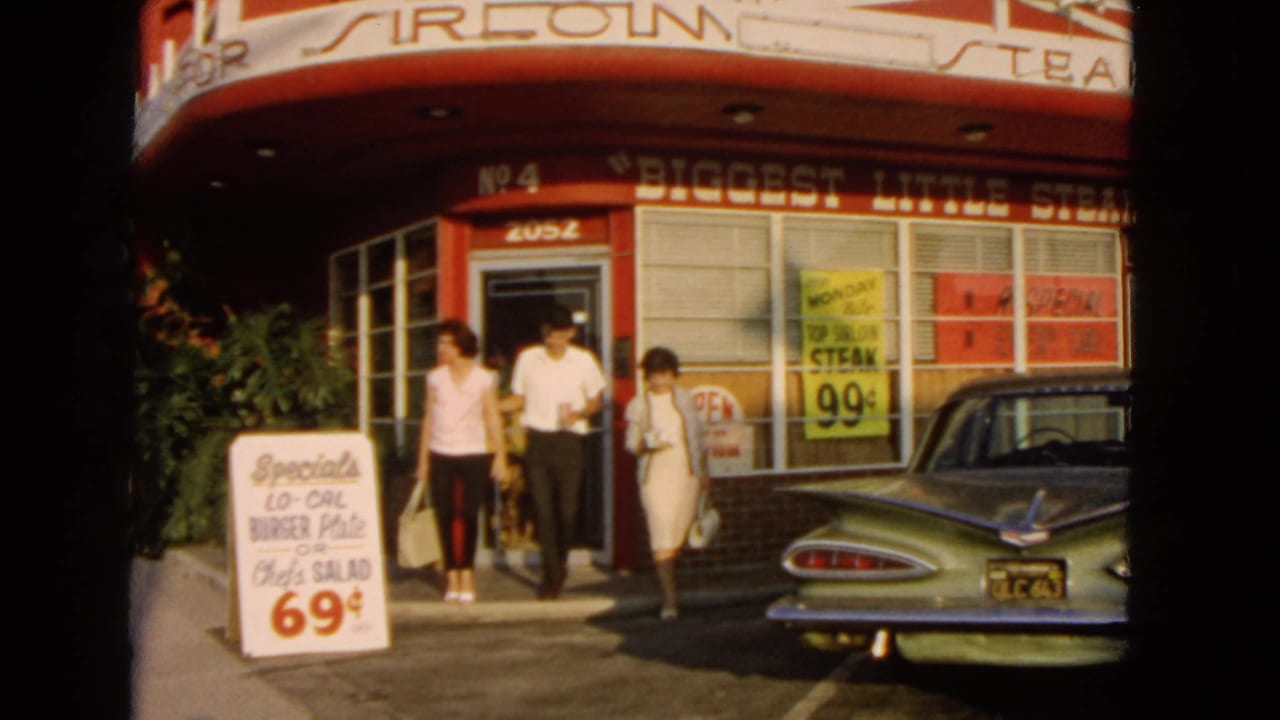 Three adults exit a red restaurant storefront beside a green tailfin car and a sidewalk special sign.