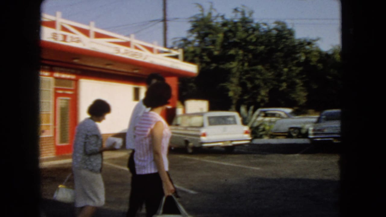 1966 San Pedro frame with adults crossing a diner parking lot near a white station wagon and other parked cars.