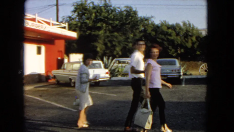 1966 San Pedro home-movie frame of three adults walking through a diner parking lot with midcentury cars behind them.