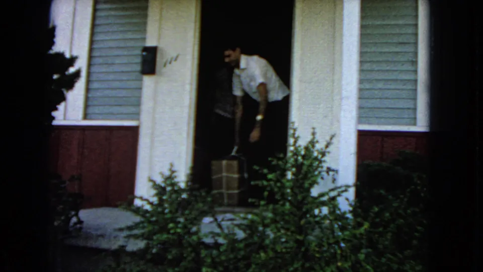 Home-movie frame showing a man stepping from a house doorway while carrying a large box, with shrubs in front.
