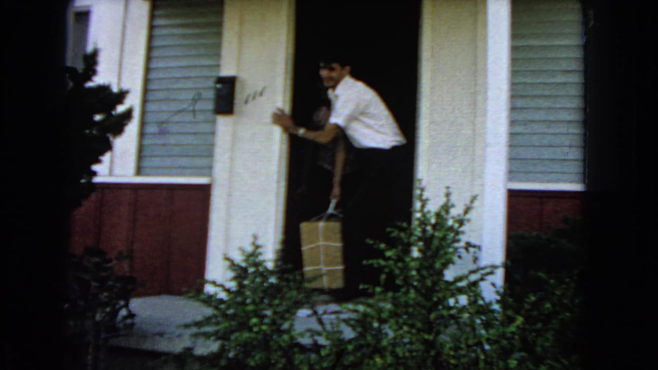 Man in a white shirt carries a wrapped package from a house doorway.