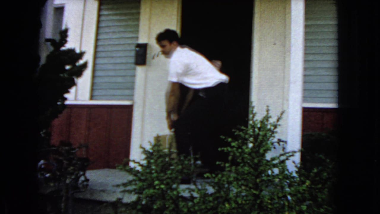 The man lowers or adjusts the wrapped package near the front porch, with house siding, a mailbox, and shrubs visible.