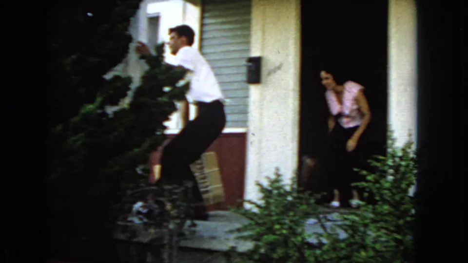 The man moves away from the doorway with the package while a woman in a patterned top watches from inside the entrance.