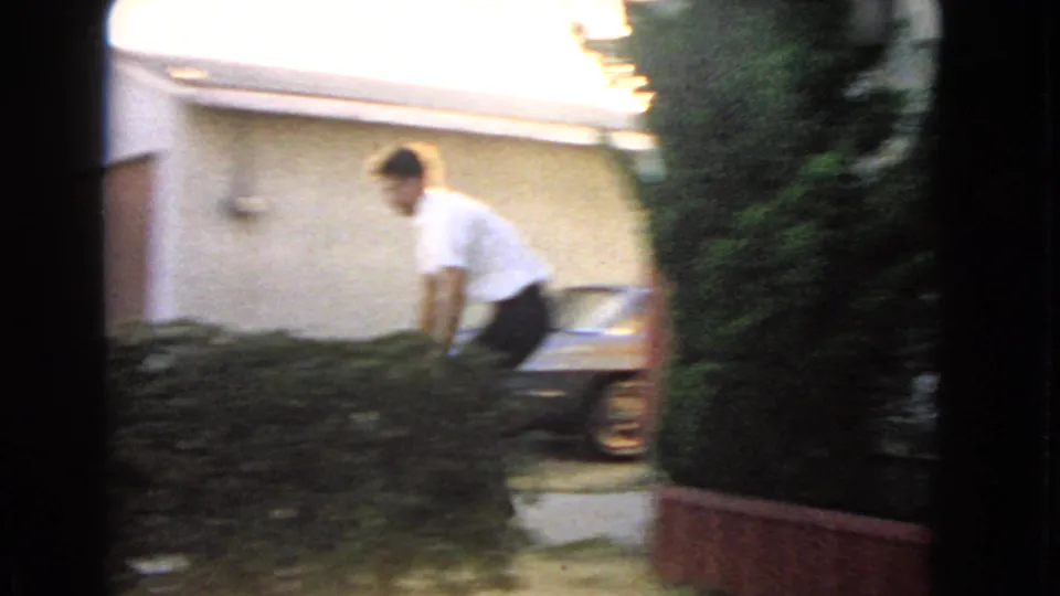 The man carries the package near a driveway or side yard, with a parked car and residential exterior visible in soft 8mm focus.