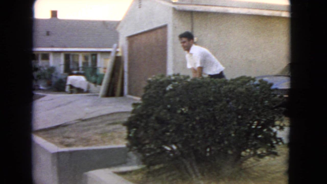 In a 1966 San Pedro home-movie frame, a man bends near a low hedge outside a suburban house.