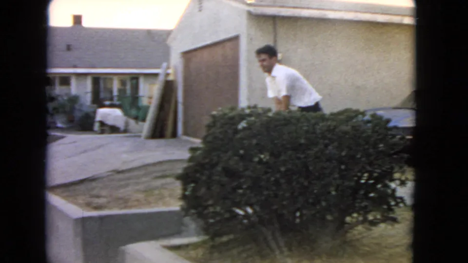In a 1966 San Pedro home-movie frame, a man bends near a low hedge outside a suburban house.