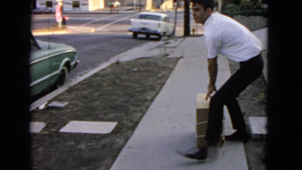 A man steadies a large package on a suburban sidewalk with vintage cars visible along the street.