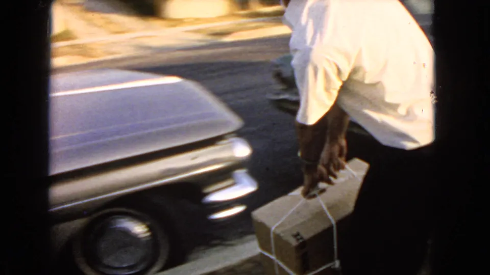 A close home-movie frame shows the man and tied package beside a vintage car at the curb.