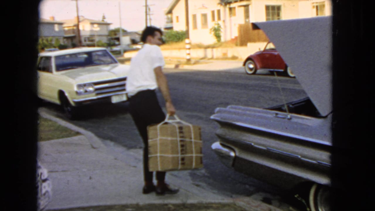 In 1966 San Pedro, a man in a white shirt carries a large wrapped box toward the open trunk of a silver car.