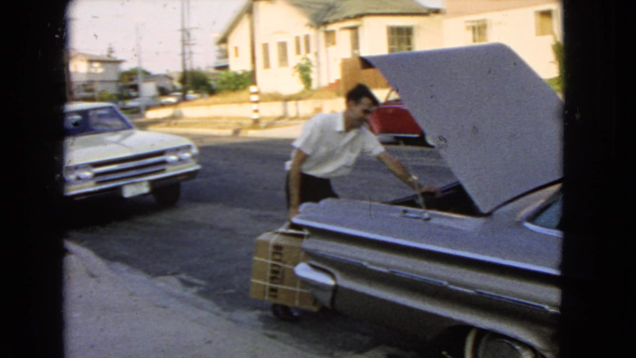Man leans toward an open car trunk while holding a large wrapped box beside the rear bumper.