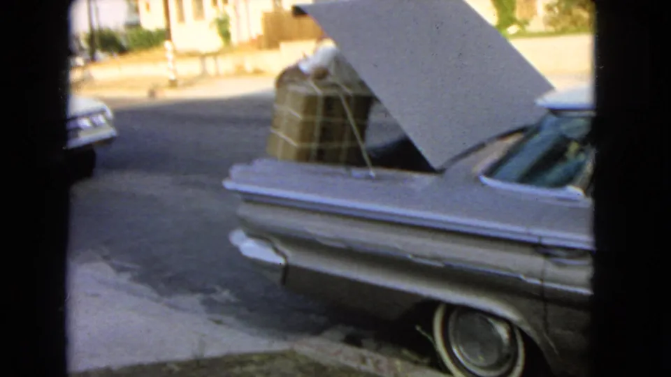 1966 San Pedro home-movie frame showing a silver sedan with its trunk open and boxes packed inside near houses.