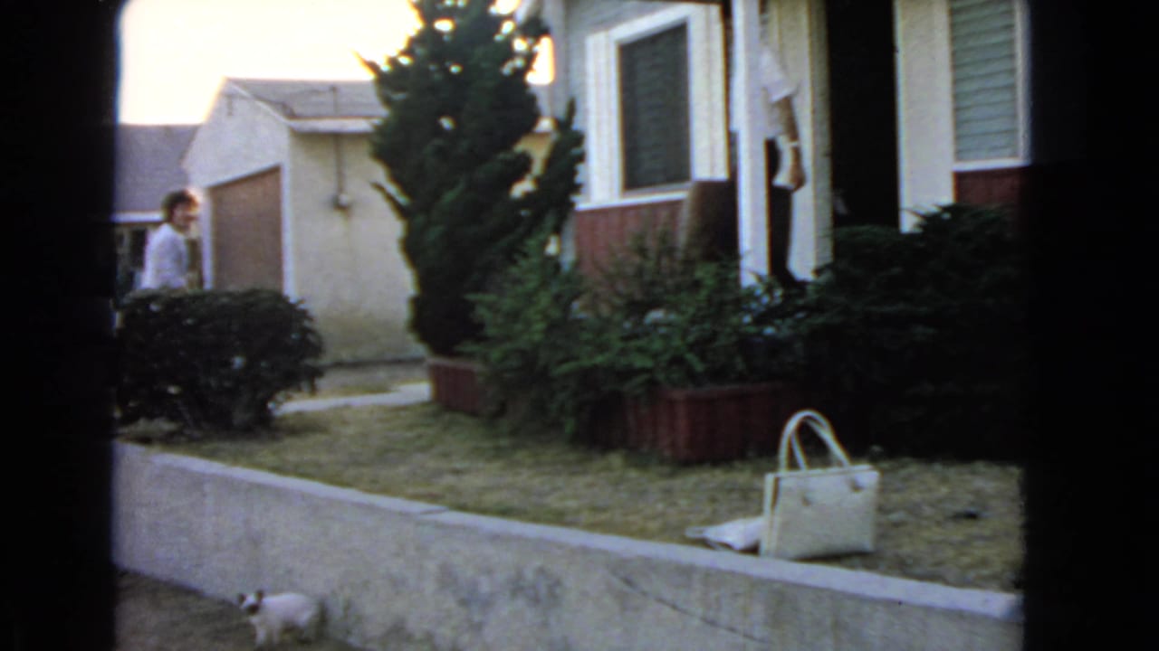 A woman stands near a San Pedro house while a white handbag rests on the lawn in this 1966 home-movie frame.